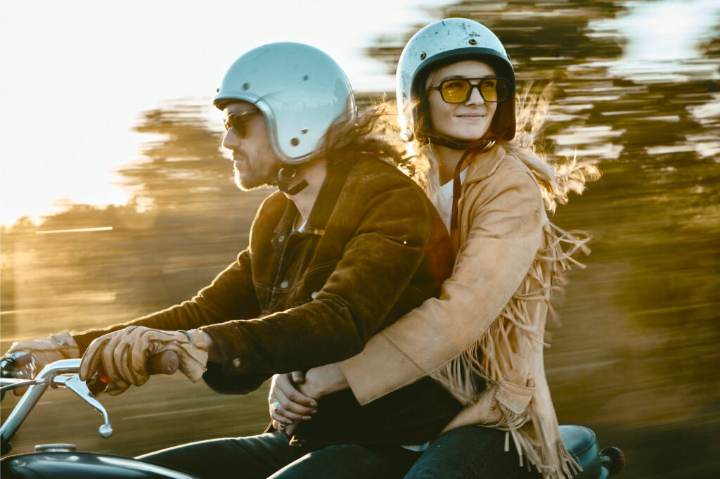 A couple rides a motorcycle wearing vintage leather jackets.