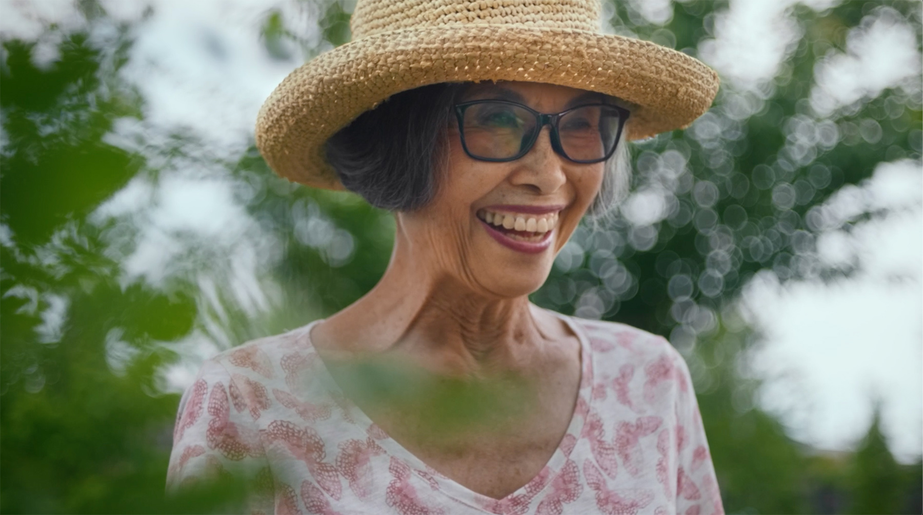 An older woman wearing a sun hat smiles in her garden.